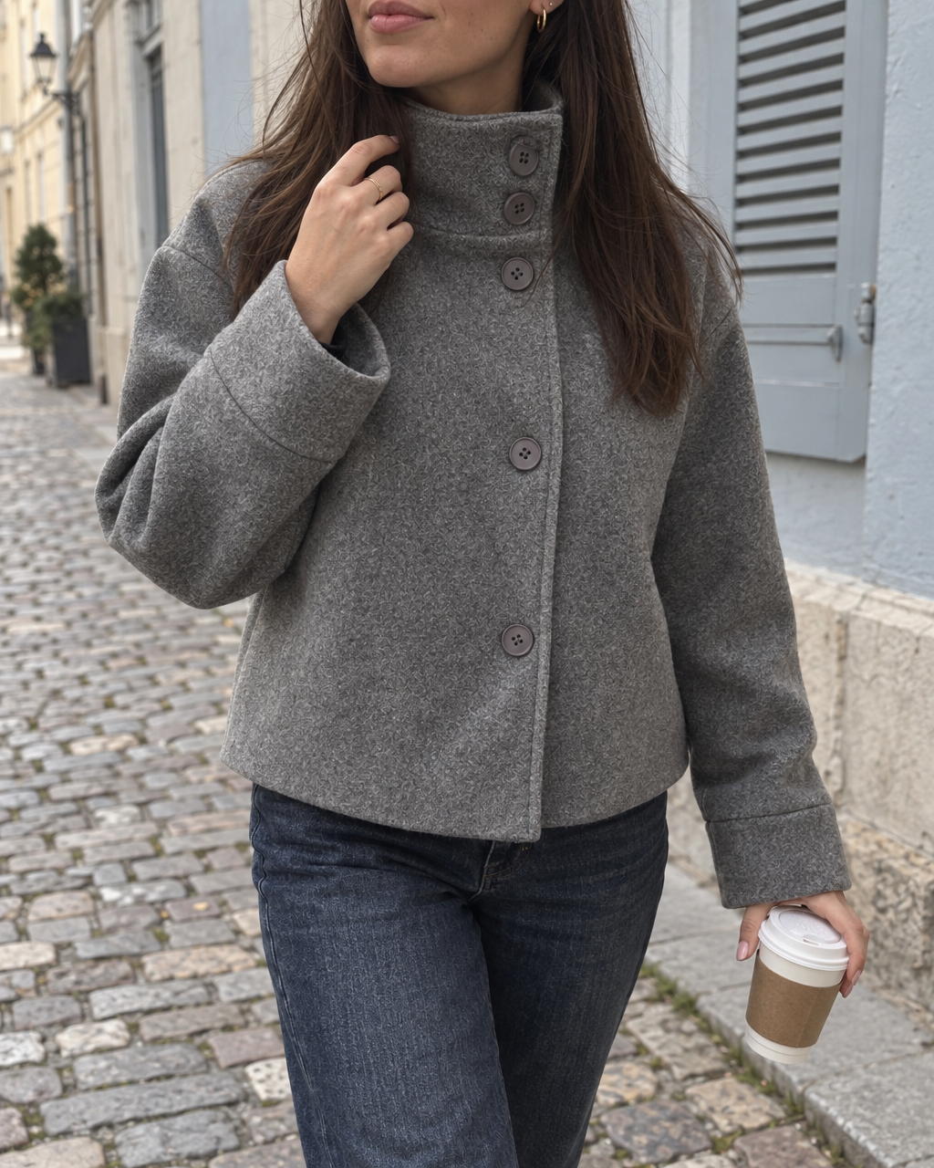 The Copenhagen Coat in Storm, hand at the stand collar, on a cobbled street with a coffee
