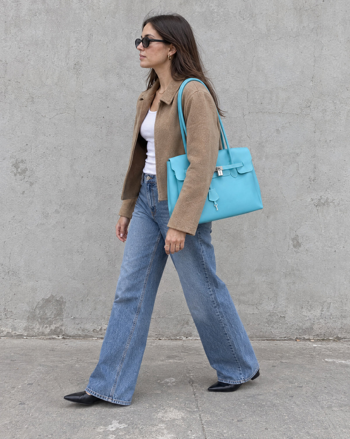 A woman in sunglasses walking past a concrete wall with the Jelly Firkin in Aqua on her shoulder