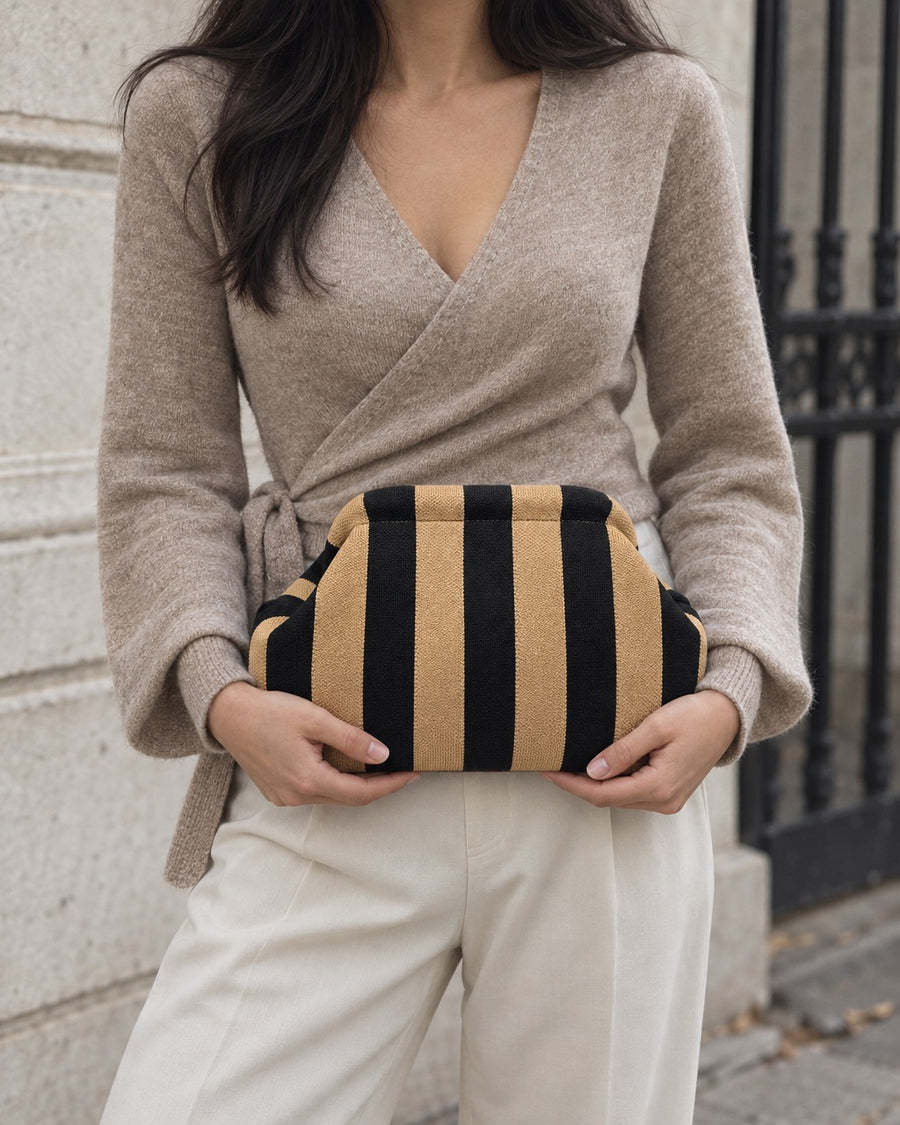 The Amalfi Clutch in Terra, a knit clutch in tan and black stripes, held at the waist by a woman in a taupe knit top and cream trousers on an old city street