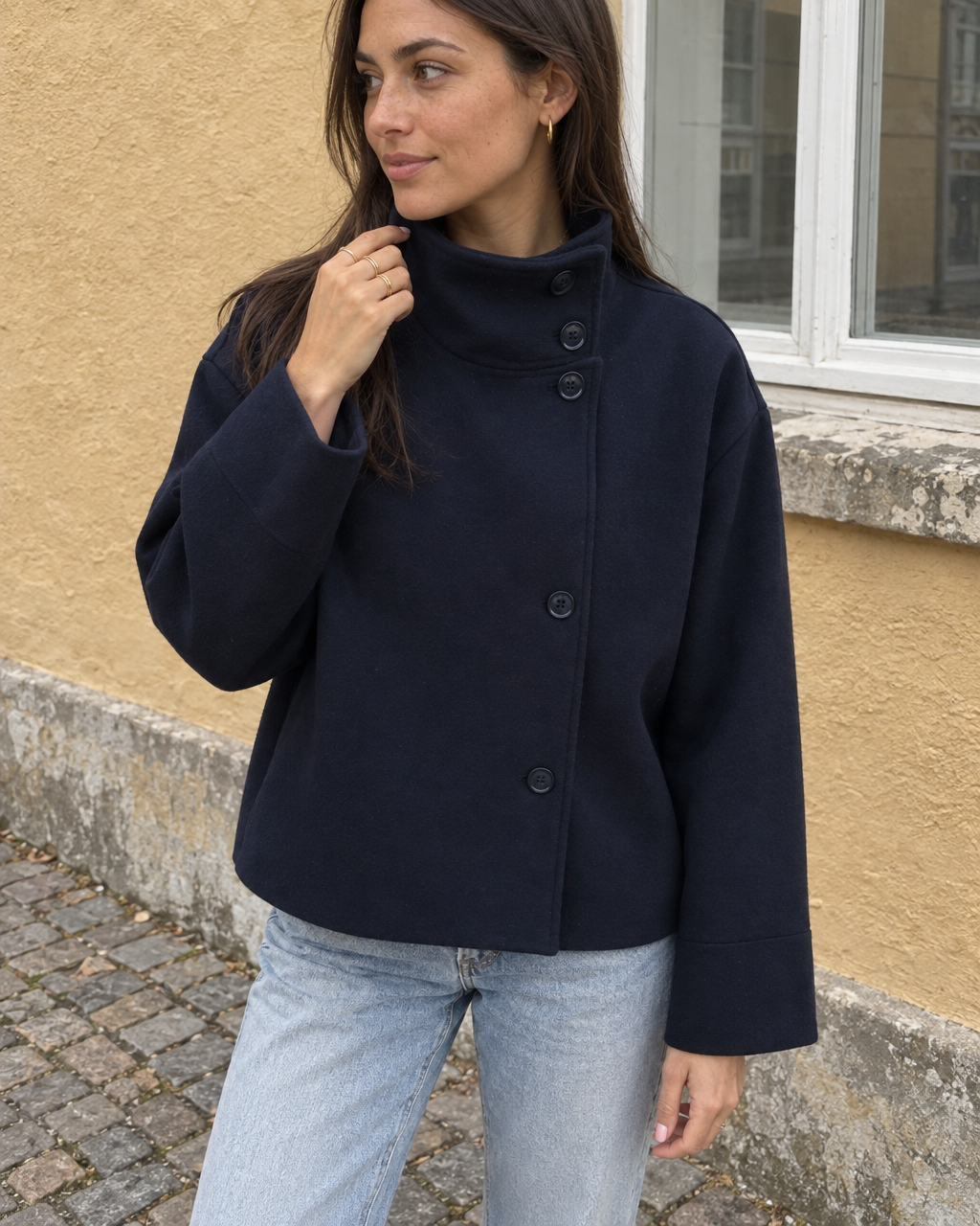 The Copenhagen Coat in Midnight, hand at the stand collar, against an ochre wall