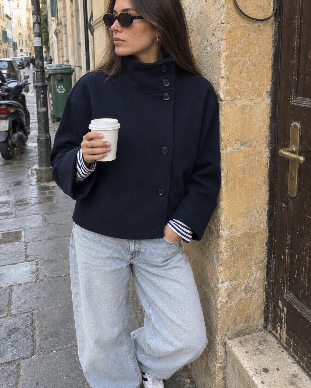 The Copenhagen Coat in Midnight on an old-town street, worn with a striped top and light jeans, holding a coffee