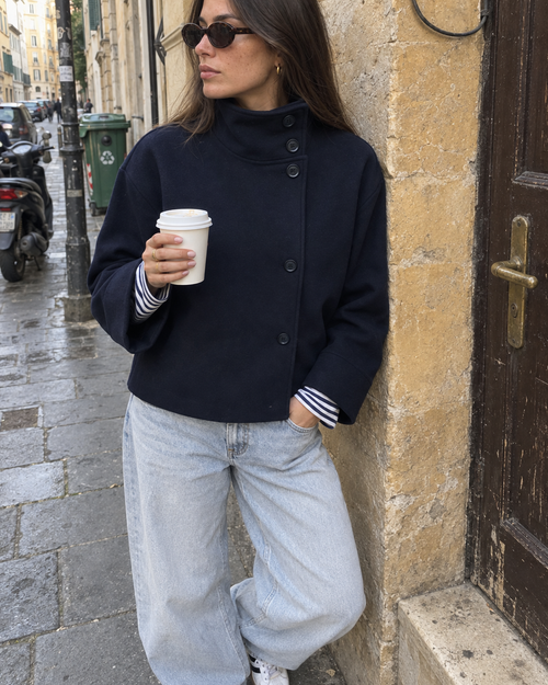 The Copenhagen Coat in Midnight on an old-town street, worn with a striped top and light jeans, holding a coffee