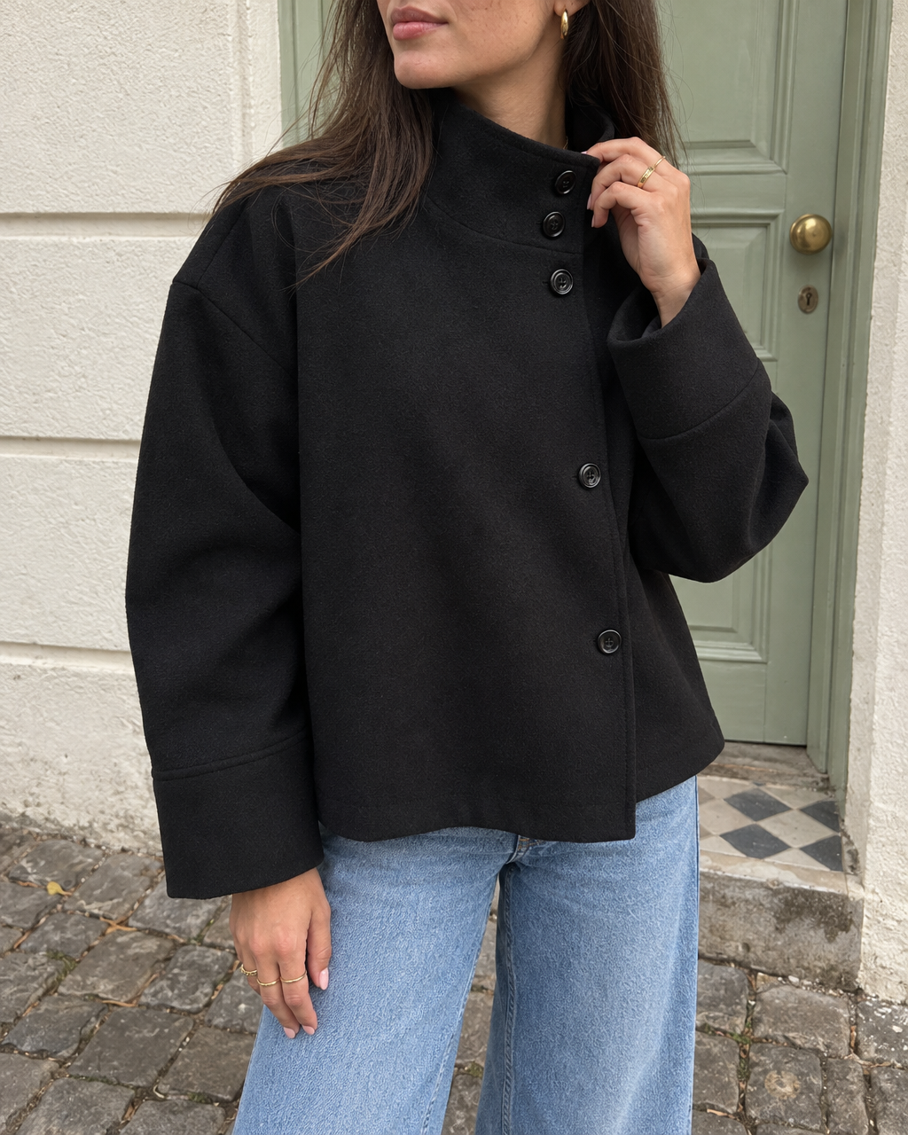 The Copenhagen Coat in Onyx, hand at the stand collar and its buttons, beside a green door